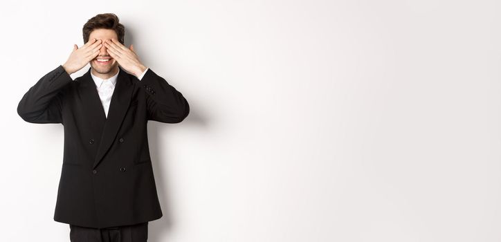 Image of handsome stylish man in black suit, waiting for christmas surprise, covering eyes with hands and smiling, anticipating presents, standing over white background.