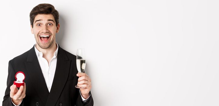 Close-up of handsome man in suit, making a proposal, giving engagement ring and raising glass of champagne, standing against white background.