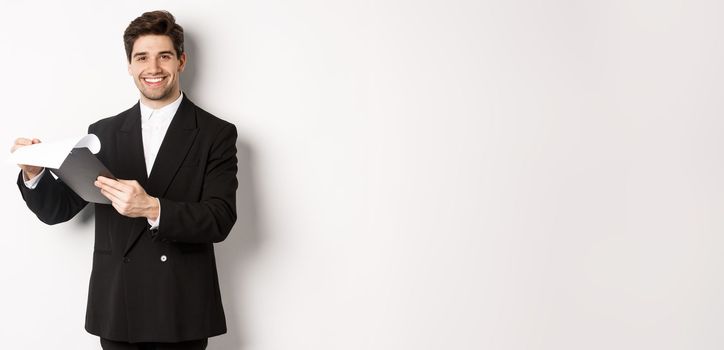 Image of confident businessman in suit looking at documents, holding clipboard and smiling pleased, standing against white background.