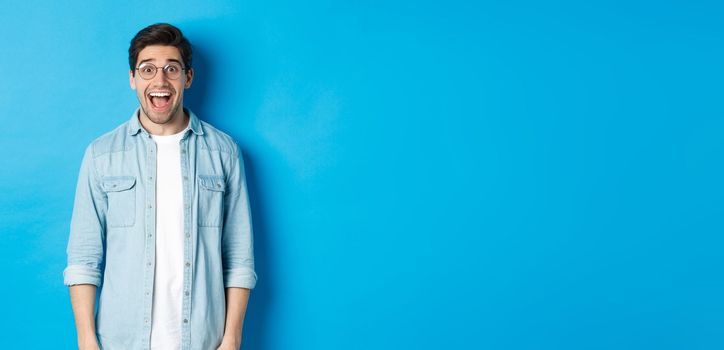 Attractive happy man in glasses looking surprised, checking out advertisement, standing over blue background.