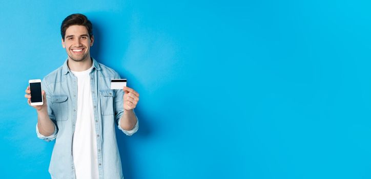 Young smiling man showing smartphone screen and credit card, concept of online shopping or banking.