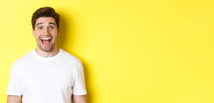 Close-up of surprised handsome guy reacting to great news, standing over yellow background in white t-shirt.