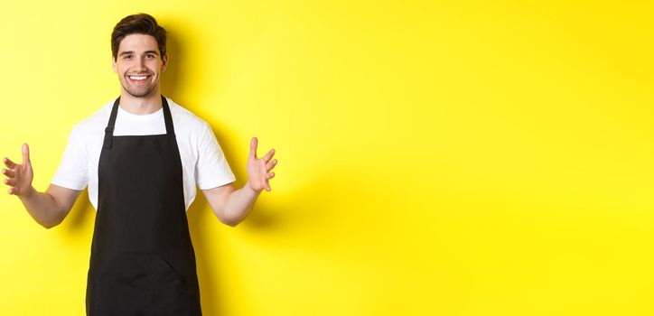 Smiling waiter in black apron holding your logo or box, spread hands as if carry something large, standing over yellow background.