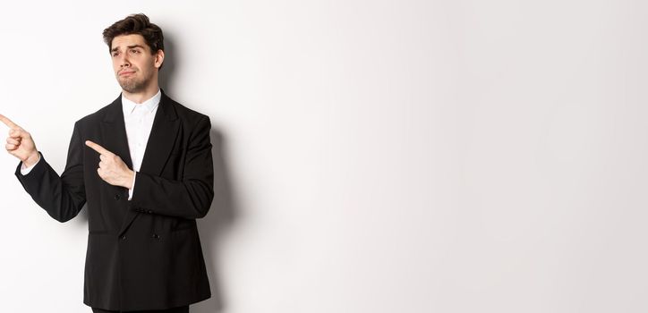 Image of upset and disappointed handsome guy in formal suit, pointing and looking left with sad face, standing over white background.