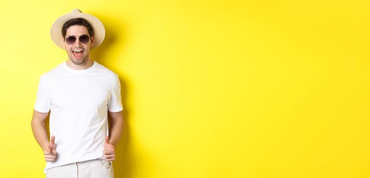 Confident and cheeky guy on vacation flirting with you, pointing finger at camera and winking, wearing summer hat with sunglasses, yellow background.