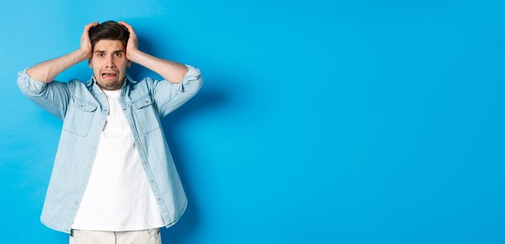 Image of man in panic holding hands on head, looking frustrated and anxious, standing against blue background.