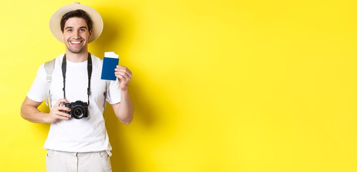 Travelling, vacation and tourism concept. Smiling man tourist holding camera, showing passport with tickets, standing over yellow background.