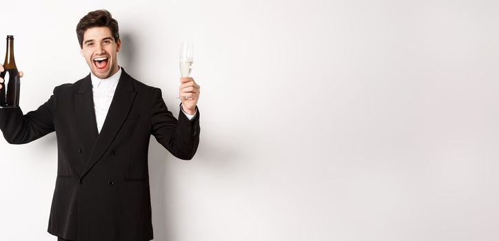 Concept of holidays, party and celebration. Handsome man in trendy suit having fun, holding bottle and glass of champagne, standing over white background.