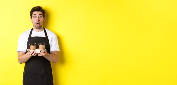 Barista serving coffee, looking surprised at camera, wearing black apron, standing against yellow background.