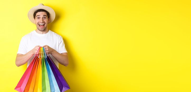 Image of happy man shopping on vacation, holding paper bags and smiling, standing against yellow background.