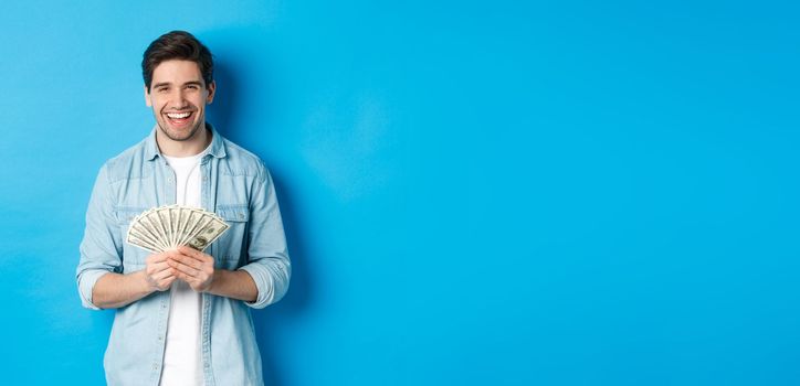 Happy successful man smiling pleased, holding money, standing over blue background.