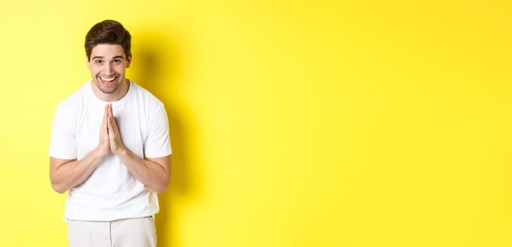 Handsome guy saying thank you, bowing and holding hands in namaste gesture, express gratitude, standing over yellow background.