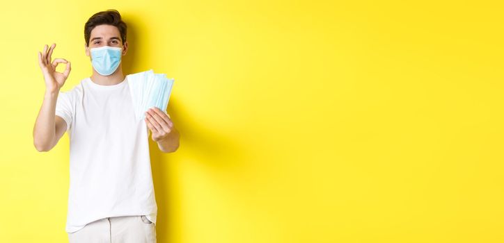 Concept of covid-19, quarantine and preventive measures. Satisfied man showing okay sign and giving medical masks, standing over yellow background.