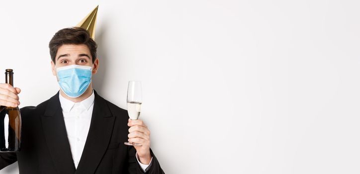 Concept of party during covid-19. Close-up of handsome man in suit, funny hat and medical mask, holding bottle of champagne, celebrating new year during coronavirus.