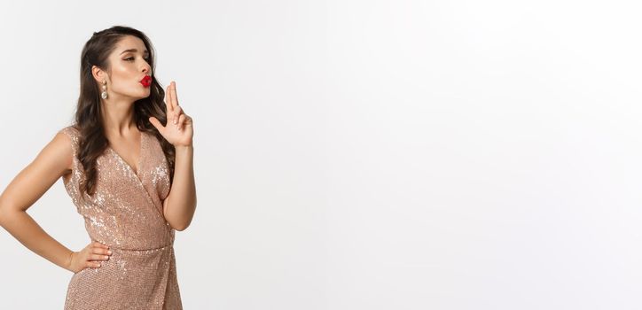 Concept of casino, celebration and party. Beautiful female model with red lips, wearing glamour dress, blowing at finger gun after making shot, standing over white background.