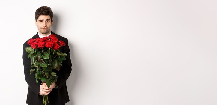 Image of handsome boyfriend in black suit, holding bouquet of red roses and smiling, being on a date, standing over white background.