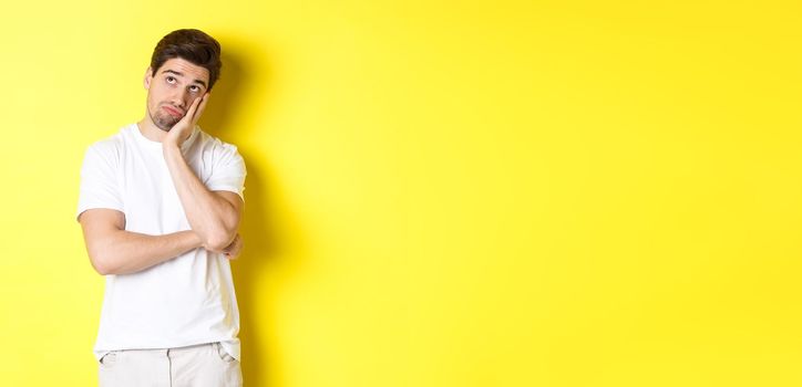 Bored and gloomy guy looking up, imaging things, standing over yellow background.