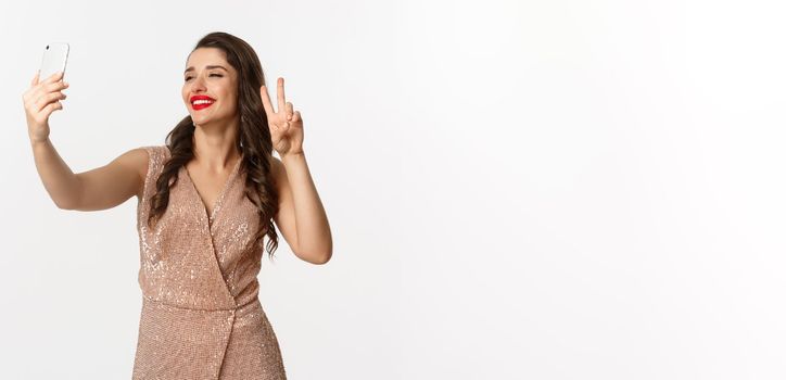 Attractive woman in luxury dress showing peace sign at phone camera, taking selfie on smartphone, posing in christmas party outfit, standing over white background.
