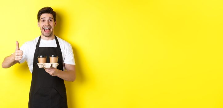 Cheerful barista recommend his cafe, holding takeaway coffee cups and showing thumbs up, standing against yellow background.