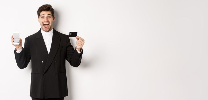 Image of handsome businessman in black suit, looking excited and showing credit card with mobile phone screen, standing against white background.