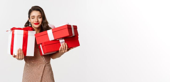 Holidays, celebration concept. Beautiful caucasian woman in elegant dress holding Christmas presents and smiling happy, standing over white background.