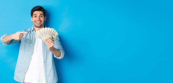 Surprised smiling man in casual clothes, pointing fingers at money, standing over blue background.