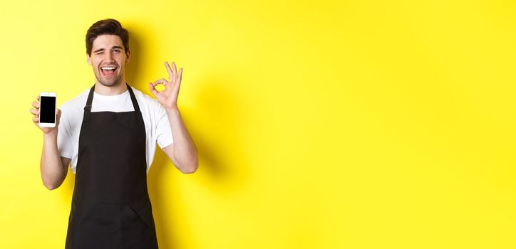 Handsome coffee shop worker showing ok sign and smartphone screen, recommending application, standing over yellow background.