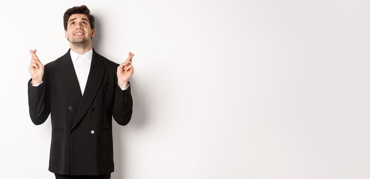 Portrait of tensed and worried handsome businessman, crossing fingers and looking up, begging god, making a wish, standing against white background in black suit.