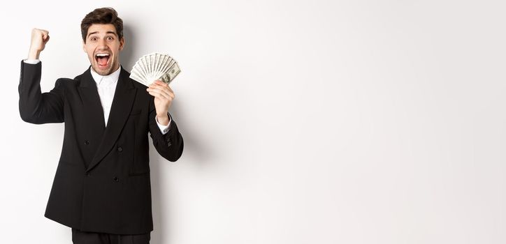 Portrait of handsome businessman in black suit, winning money and rejoicing, raising hand up with excitement, standing against white background.