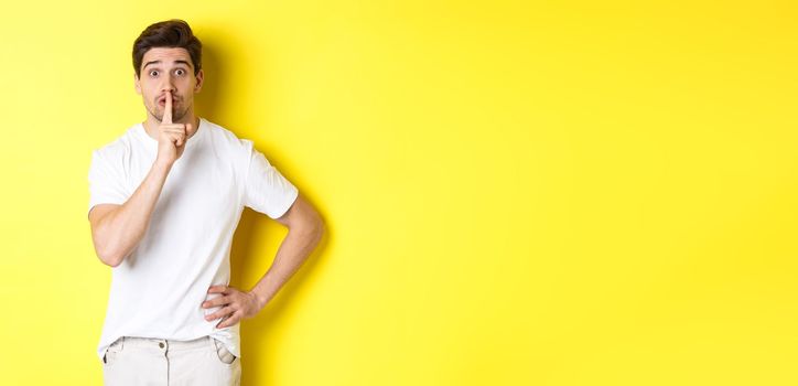 Image of excited guy planning surprise, shushing at camera, press finger to lip in hush sign, standing over yellow background.