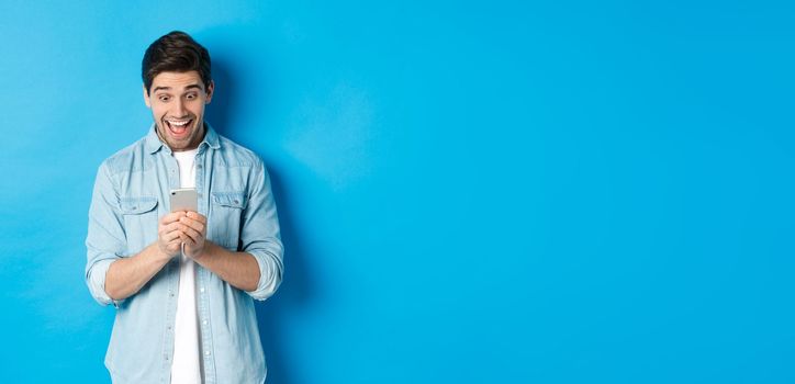 Image of excited man smiling while looking at mobile phone, shopping online on smartphone, standing against blue background.