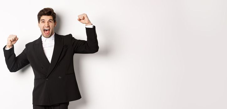 Portrait of successful businessman in black suit, become champion, raising hands up and shouting yes, triumphing and celebrating victory, standing against white background.