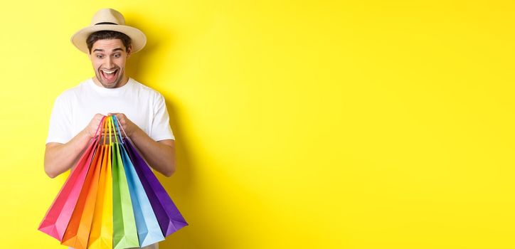 Image of happy man shopping on vacation, holding paper bags and smiling, standing against yellow background.