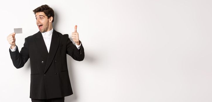 Image of excited handsome businessman, showing credit card and thumb-up, standing against white background.