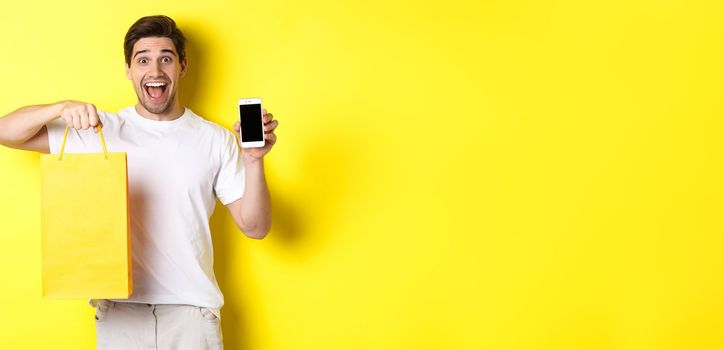 Concept of mobile banking and cashback. Young happy guy holding shopping bag and showing smartphone screen, yellow background.