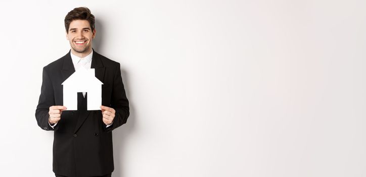 Image of handsome real estate agent in black suit showing home maket, smiling at camera, standing against white background.