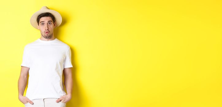 Confused guy traveller in straw hat looking puzzled, stare at camera, standing over yellow background. Copy space