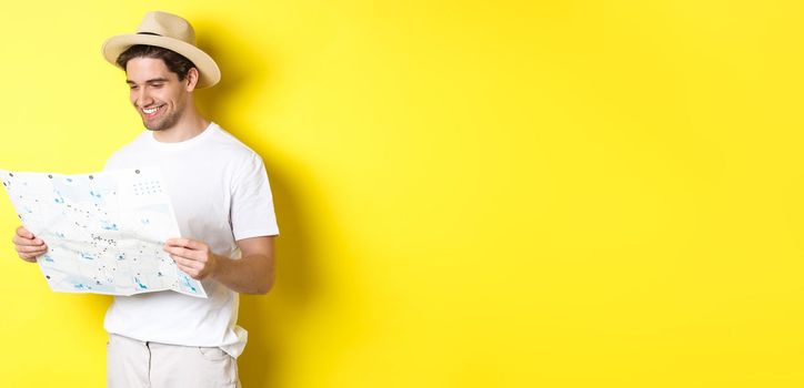 Travelling, vacation and tourism concept. Smiling happy tourist looking at map with sightseeings, standing against yellow background.