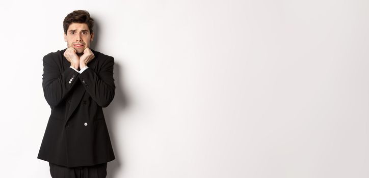 Image of scared and insecure young businessman in suit, trembling from fear and looking horrified, standing over white background.