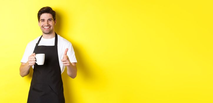 Barista bringing coffee and pointing finger gun at camera, standing in black apron against yellow background.