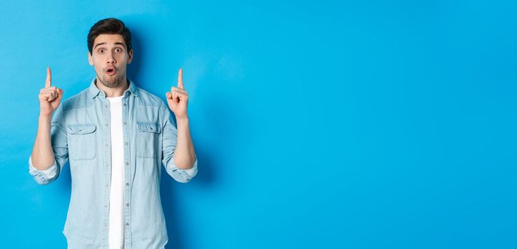 Portrait of amazed handsome man pointing fingers up, showing logo on blue background.