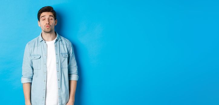 Handsome man waiting for kiss, pucker lips and close eyes while standing against blue background.