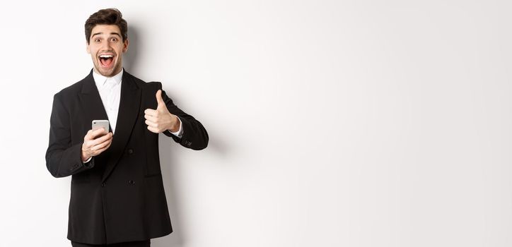 Portrait of handsome man in trendy suit showing thumbs-up in approval, using mobile phone app, smiling pleased, standing over white background.