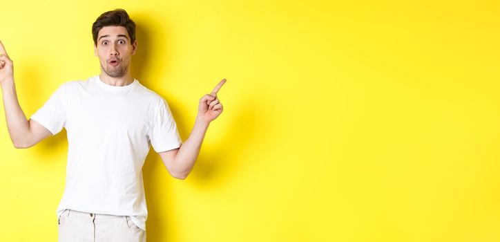 Handsome man pointing fingers sideways, showing two promos, standing over yellow background.