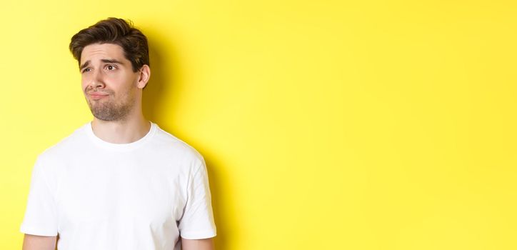 Reluctant guy in white t-shirt looking left, grimacing skeptical and displeased, standing over yellow background.