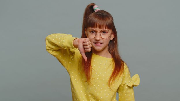 Dislike. Upset young preteen child girl kid showing thumbs down sign gesture, expressing discontent, disapproval, dissatisfied bad work indoors. Displeased toddler children on gray studio background