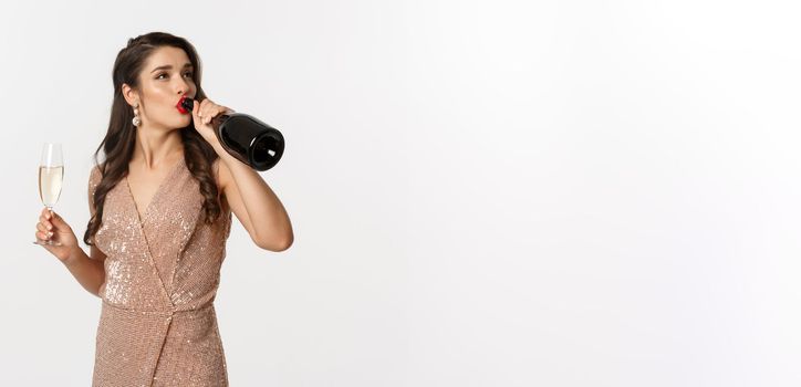 Winter holidays celebration concept. Careless woman in elegant dress drinking champagne from bottle and looking unbothered, standing over white background.