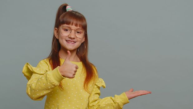 Happy toddler girl showing thumbs up pointing empty place, advertising area for commercial text, copy space for goods promotion banner, copy-space. Young child kid. Studio shot on gray wall background