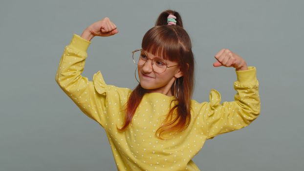 I am strong and healthy. Young toddler girl in glasses showing biceps and looking confident feeling power strength to fight for rights, energy to gain success win. Preteen child kid on gray background
