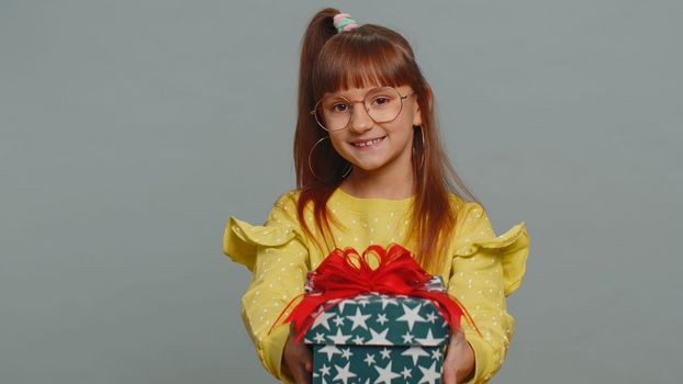 Lovely smiling young preteen child girl kid presenting birthday gift box stretches out hands, offer wrapped present career bonus, celebrating party. Little toddler children on studio gray background